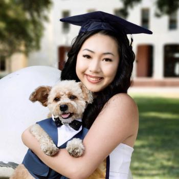 Renee is centered in the middle of the frame, facing the camera and smiling. She is carrying a fluffy, white dog. The dog is dressed in a tuxedo and bowtie. Renee is wearing a white dress and a graduation cap. 