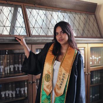 Emily Choto poses for the camera while holding onto a bookshelf. She is wearing graduation regalia from USF.