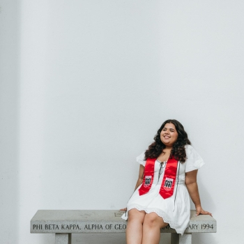 NHC member Ann Sunny wearing a white dress and graduation stole, sitting on a bench.