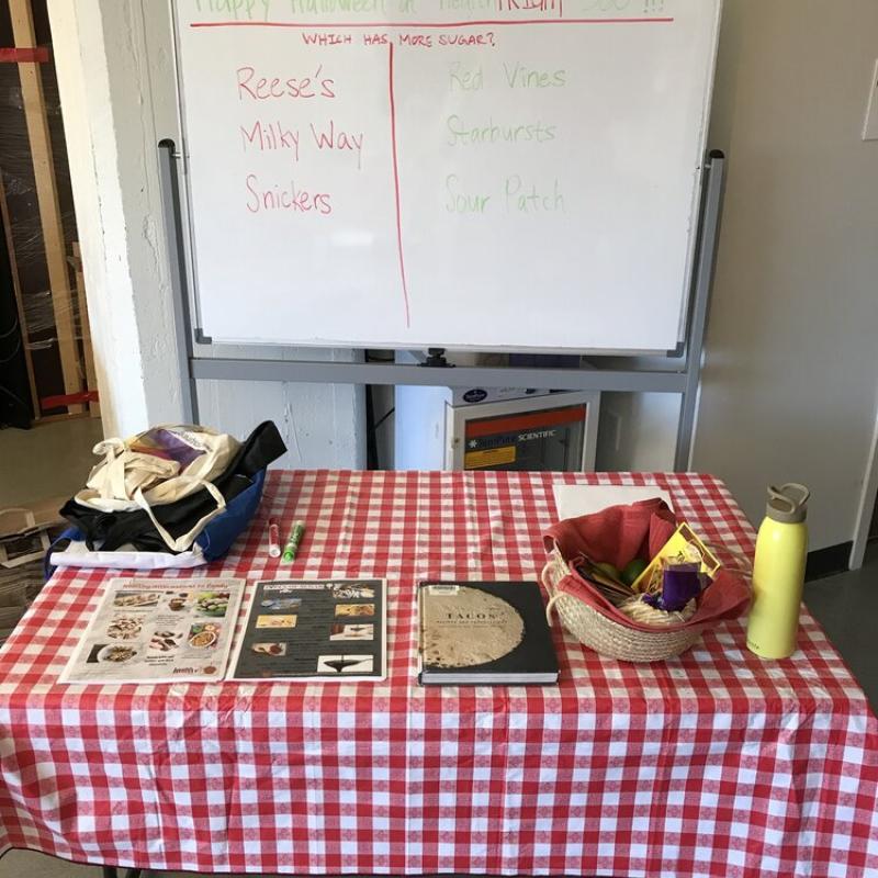 A table at HR 360 Food Pharmacy with a few recipe books, a basket, and a whiteboard behind the table reading "Happy Halloween at HR 360!" and a poll asking which candy has more sugar (Reese's, Milky Way, Snickers, Sour Patch, Starbursts, Red Vines). 