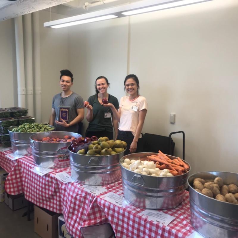 NHC SF AmeriCorps Members (From left to right, Wesley, Julia, Kristin) standing at the HealthRIGHT 360 Food Pharmacy table, with bowls of fruits and vegetables set up on top of the table. 