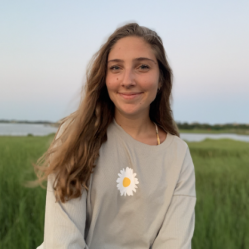 NHC PGH member Miranda soft-smiling in a field with a daisy on her sweatshirt