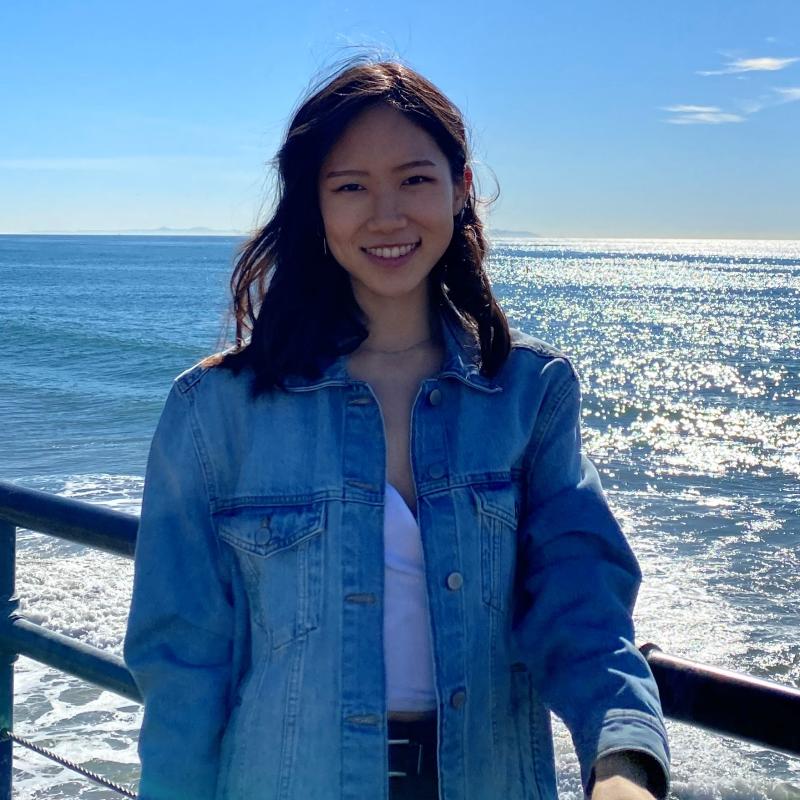 NHC SF AmeriCorps Member Clara Chung standing with a background at a pier. Ocean waves and shimmering sunlight frame her.