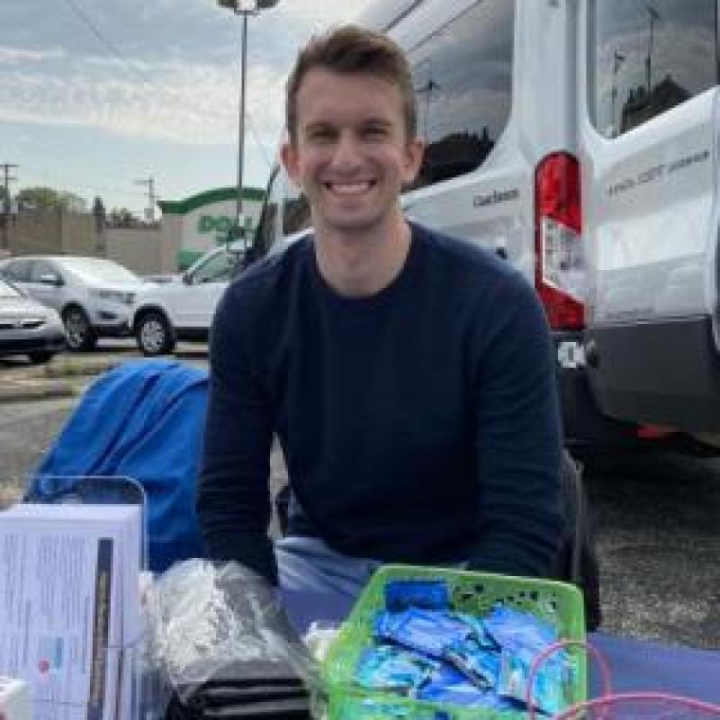 NHC PGH member Brett smiling at a health event outside, behind a table of health education materials