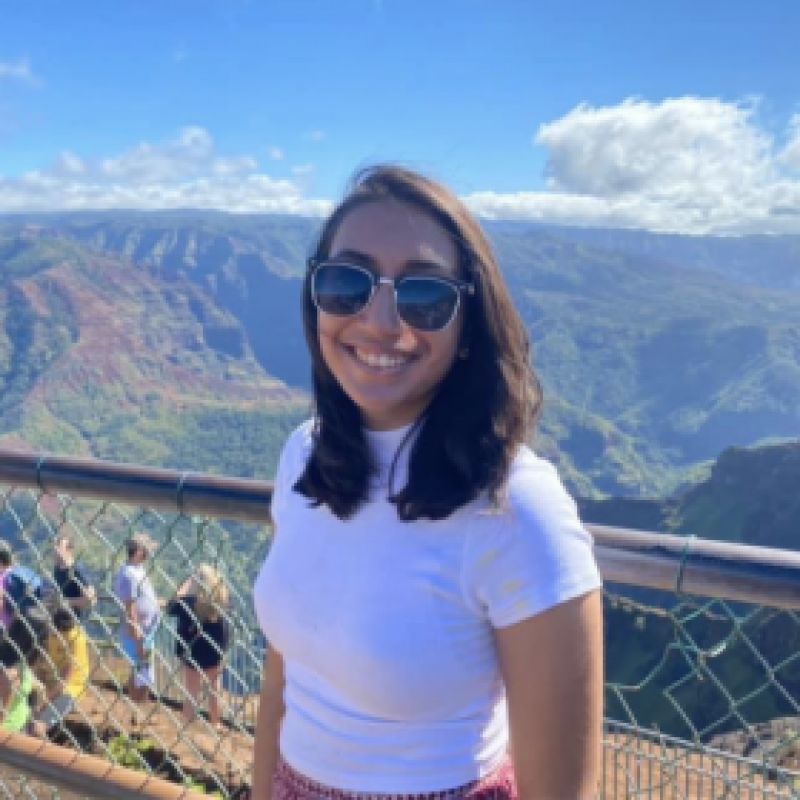 NHC PGH member Chinmayi smiling on a sunny day with a scenic mountainous background wearing sunglasses
