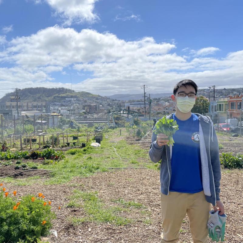 NHC SF member Jacky Chu holding a plant in their right hand. The background is a garden with semi-cloudy blue skies overhead. 