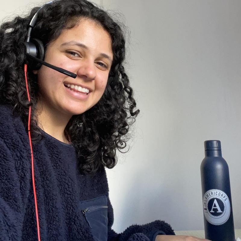 NHC SF member Saba Moussavian serving from home, wearing the audio headgear and microphone. An AmeriCorps water bottle is seen in the background against a white background.