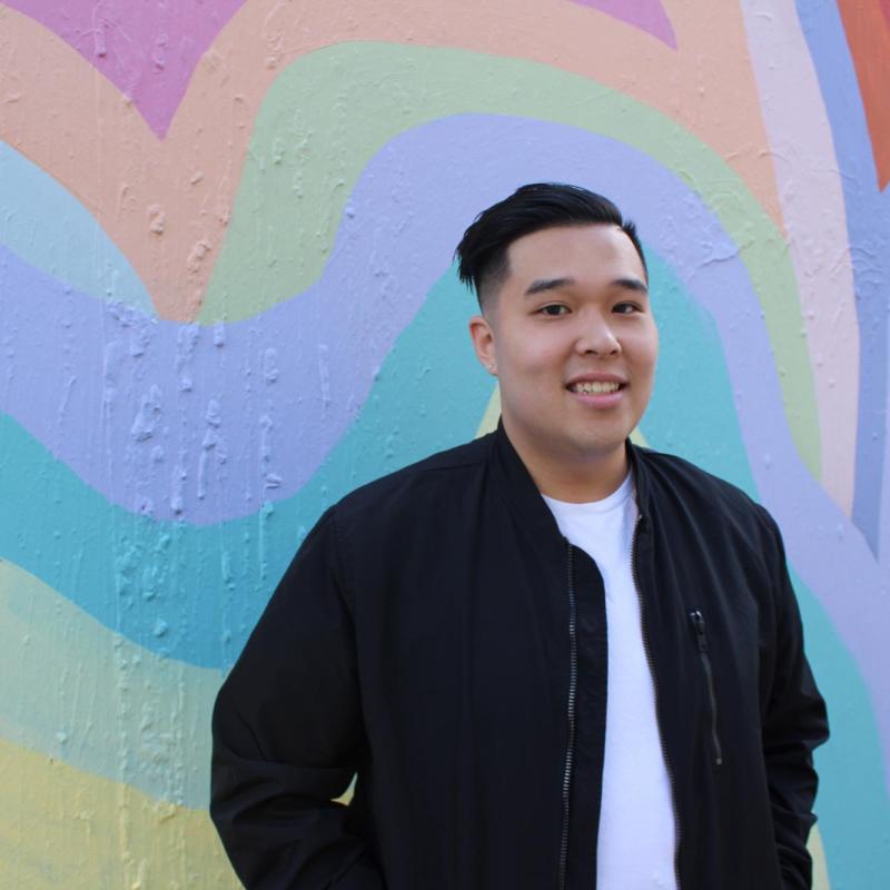 NHC SF member Antony Nguyen, smiling, wearing a black jacket over a white t-shirt. The background is a mural of overlapping, colorful painted hearts, next to a drawn symbol.