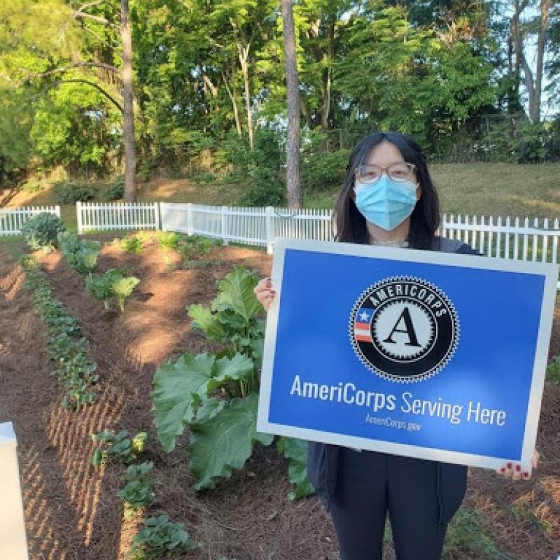 Jennifer standing in a garden and holding a sign that reads "AmeriCorps Serving Here"