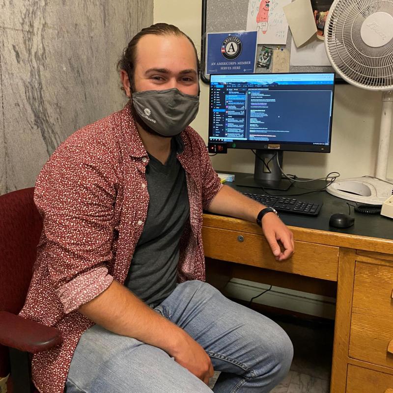 Eli Solomon slightly turned to the right, facing the camera. Eli is wearing a face covering, an open button down shirt with a grey undershirt, and lightwash jeans. He is sitting facing away from the computer and desk. 