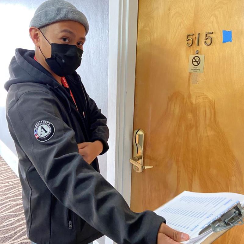 Jeffrey Levita facing the camera with a facemask on. He is extending his right arm out to the lower right corner of the frame while holding a clipboard. On his right shoulder is the AmeriCorps logo.
