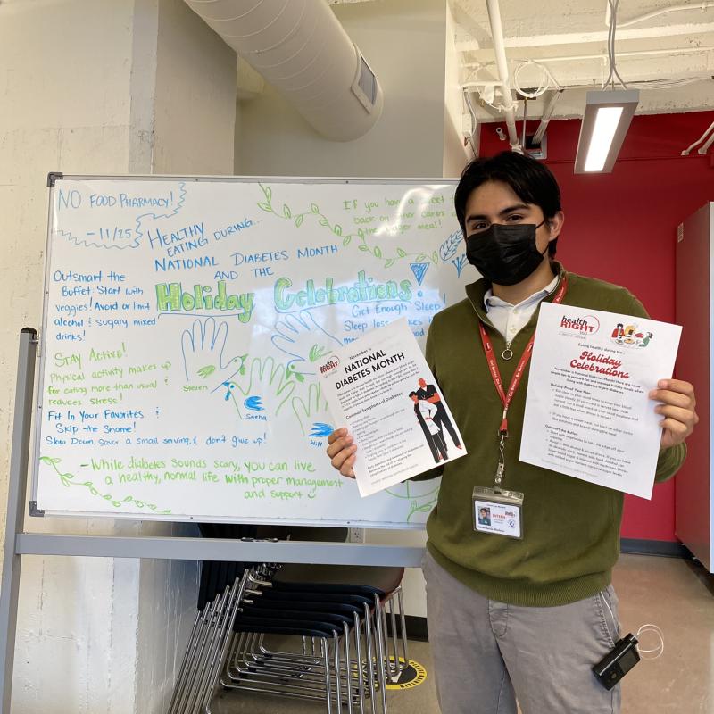 Steven is seen here facing the camera, holding up two educational material flyers regarding nutrition and diabetes care awareness. To his right is a white board with writing at the center about healthy eating during the holiday months for patients with diabetes.