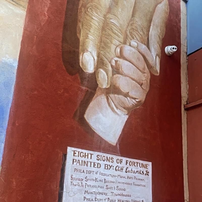Mural of an adult hand being grasped by a childs hand on a red wall. Under the image is text, some illegible, but the last line is a quote that reads "my humanity is bound up in yours, for we can only be human together" - Desmond Tutu