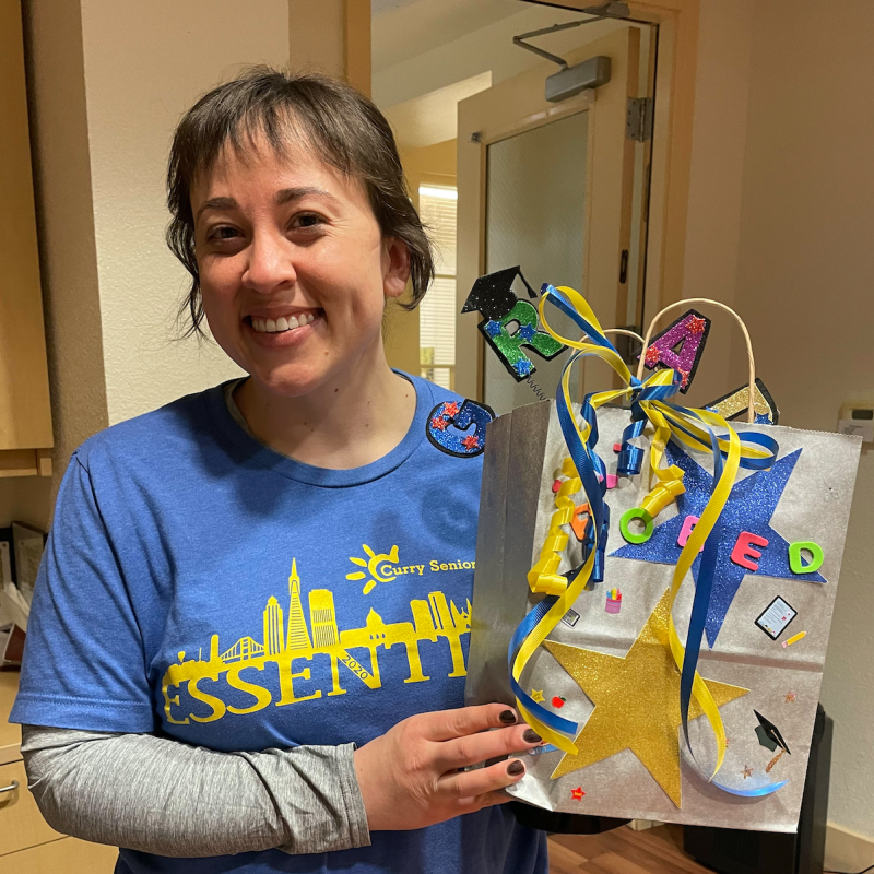 Hannah Griggs is seen here smiling towards the camera. She is holding a gift bag with ribbons of blue and yellow colors coming over the front of the bag. There is a yellow star on the bottom left front facing corner of the bag, and an adornment with a black graduation cap protruding from the top of the bag. 
