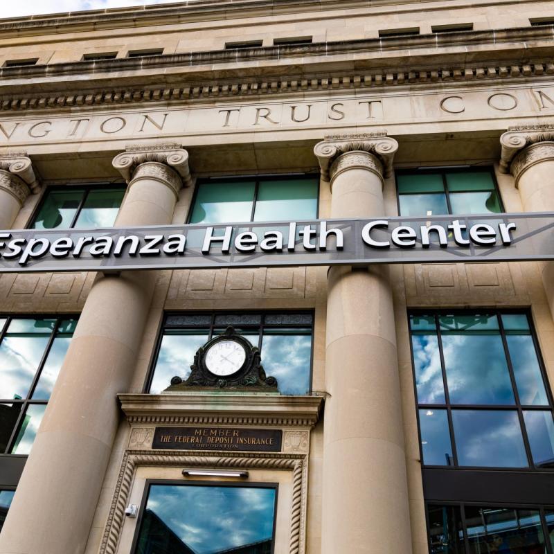 Looking up at a building with tan columns. There is an ornate clock over the front door and above that a sign that says "Esperanza Health Center"