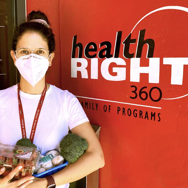 Mariana Carranza is seen standing in front of a HealthRIGHT 360 sign. She is holding assorted produce to give to patients. 