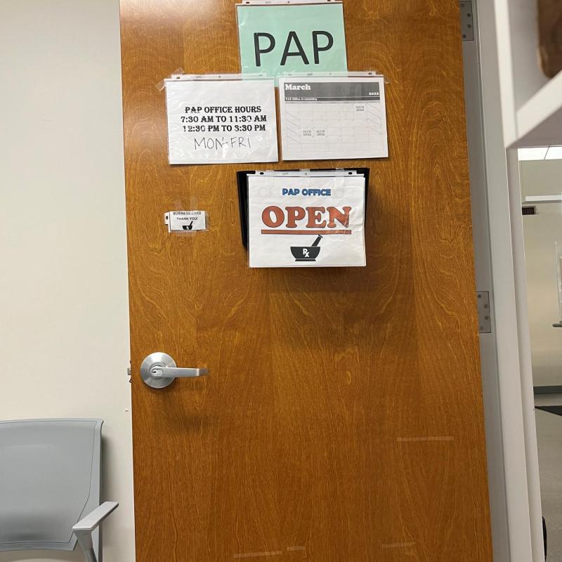 An open door with four signs hanging. At the top a sign reads "PAP" under that are two, on the left lists the office hours and on the right is a calendar showing March. The last sign is below all of them and it reads "PAP Office Open" 