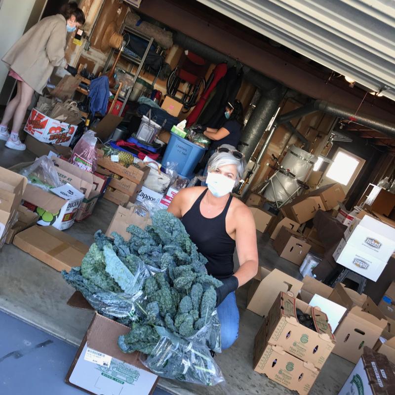 Janna is seen in the center of the frame, facing the camera with a mask on. She is squatting behind a box that contains kale, a green leafy vegetable to hand out to patients.