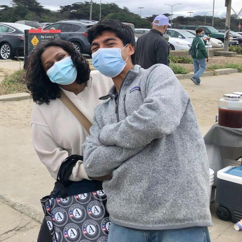 Two people are in the center of the photo. Johnny Reyes (right) and Sneha Suresh (left) are leaning into each other, arms crossed in front of their bodies. They both have a facemask on.