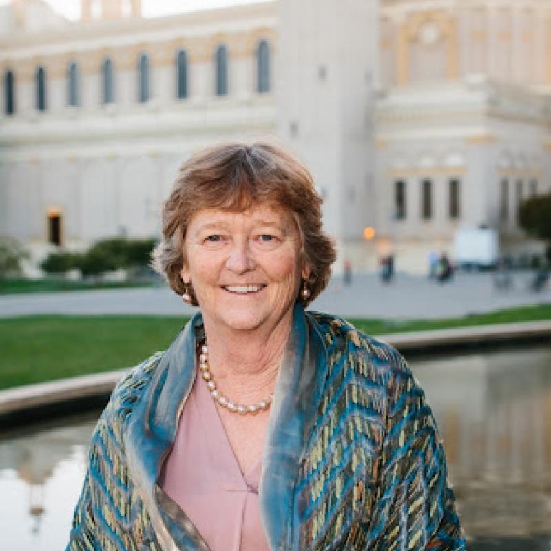 A person is standing at the center of the photo and is seen from the waist up. They have their arms crossed in front of them and are smiling at the camera. In the background there is a tall building.