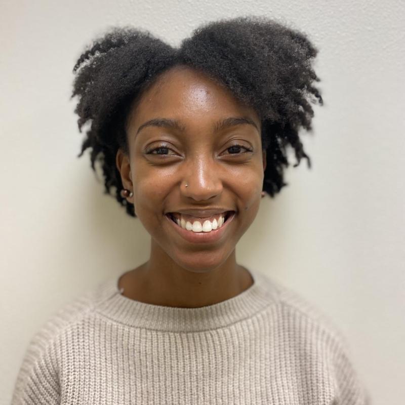 A person (Imani) is in the center of the frame smiling at the camera. She is wearing a knitted sweater. She is standing against a blank background.
