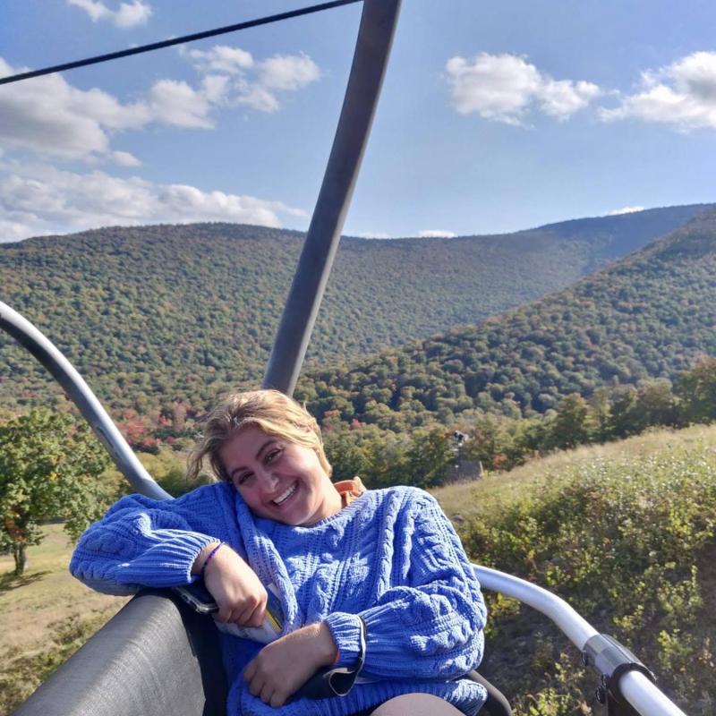 Sarah wearing a blue sweater sitting on a ski-lift during the summer, there are hills covered in green trees behind her.