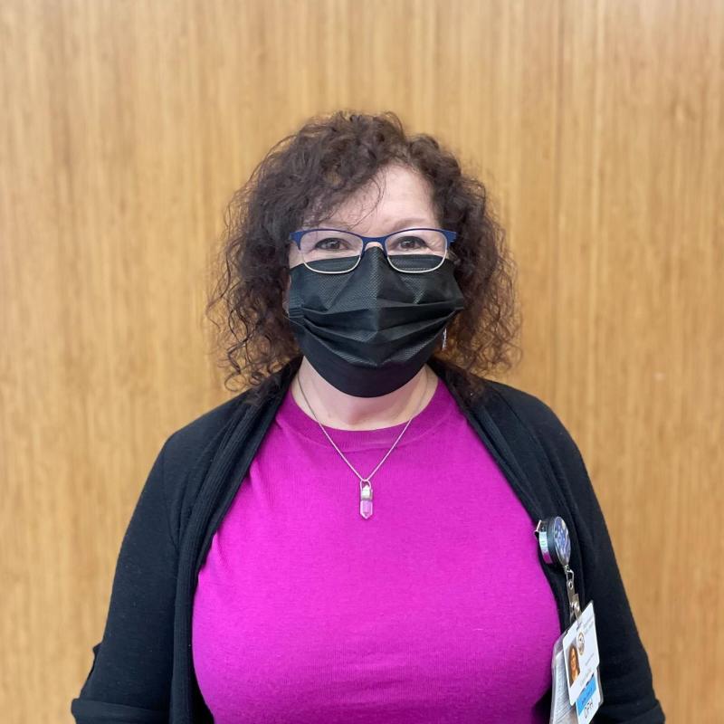 Person in middle of screen is standing facing the camera. Person is wearing a facemask and an open cardigan over a magenta long-sleeve shirt. 