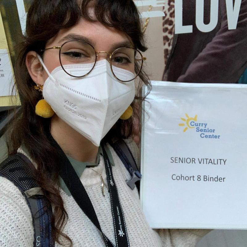 Sarah is facing the camera. She is holding a binder that reads "Curry Senior Center; Senior Vitality; Cohort 8 Binder". 