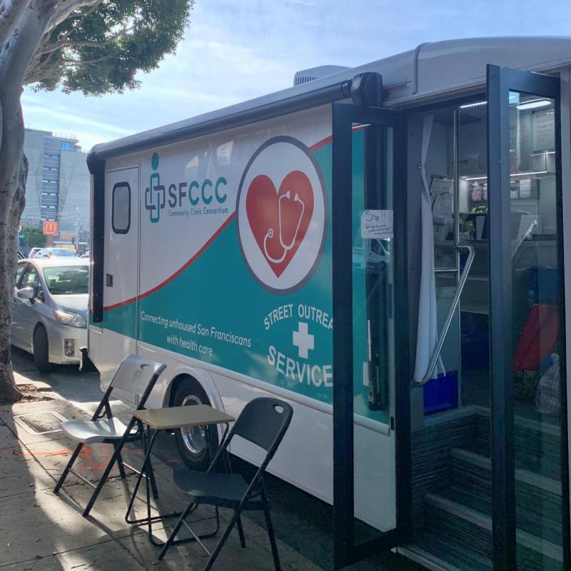 A photo of the Street Outreach Services (SOS) van, parked. There's a table with two folding chairs on either side of it. On the van, the sign reads "Connecting unhoused San Franciscans with health care."