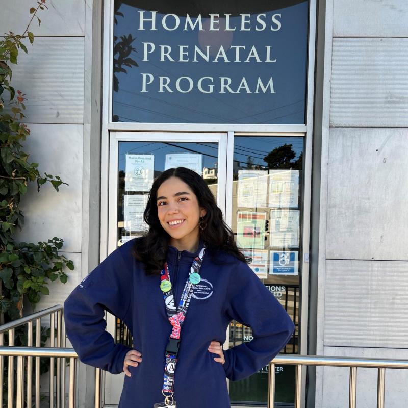 Jasmine is standing in the middle of the frame in front of the entrance to a building. Behind her and above the door is a sign that reads Homeless Prenatal Program. 