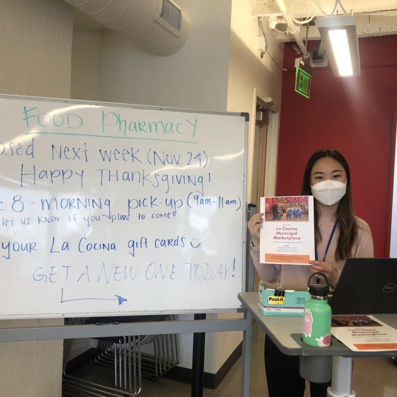 Joelle is holding a flyer for La Cocina's Municipal Marketplace. Food Pharmacy's whiteboard indicating information regarding Thanksgiving closure, a December food pharmacy pickup, and La Cocina giftcards is to her left.