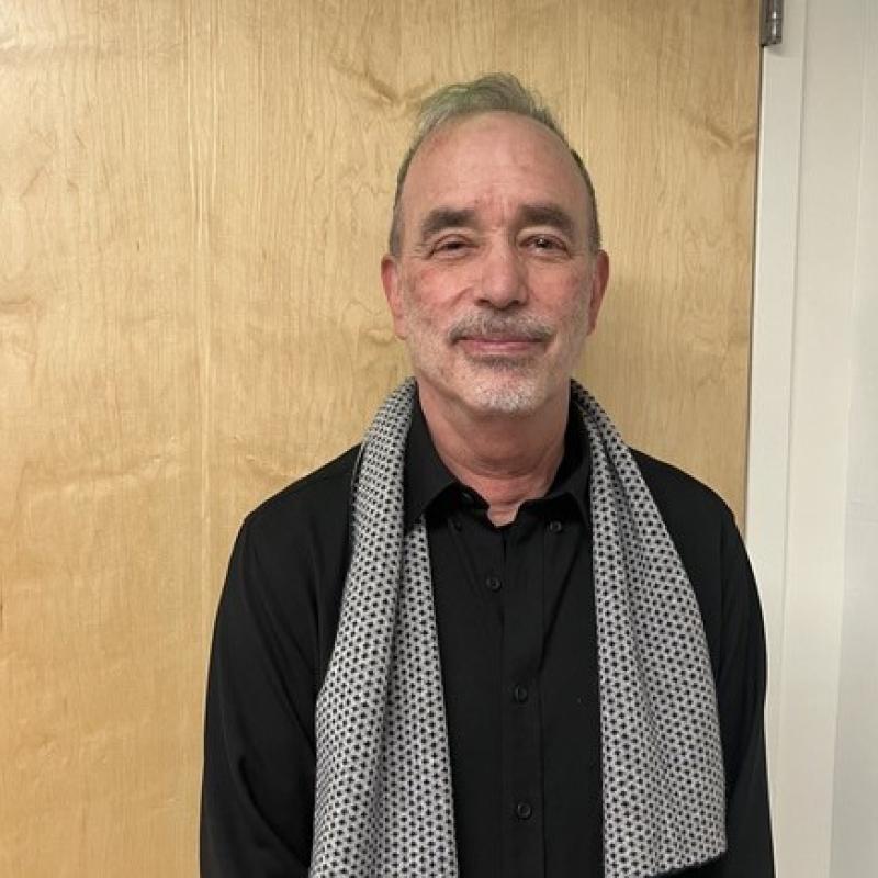 Michael is standing in front of the camera. They are wearing a black button down shirt and a grey scarf. 