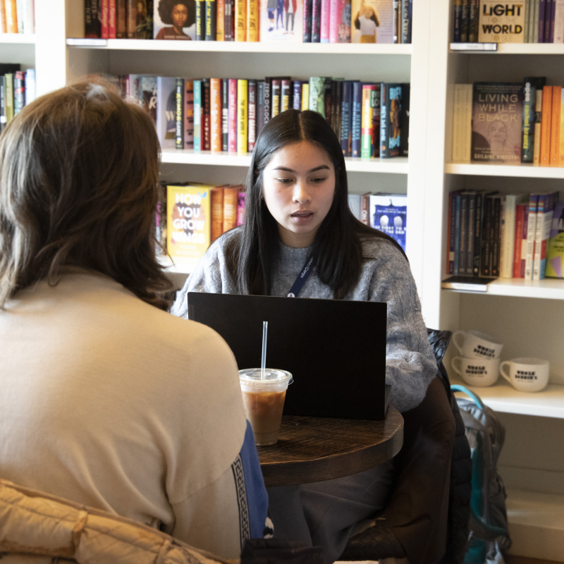 Patricia sitting in front of a bookshelf with her laptop. There is another person sitting across from her whose face we cannot see.