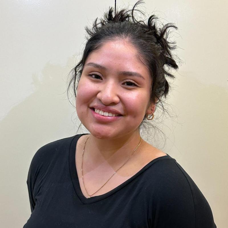 Paola Zavala is standing in the middle of the frame, facing the camera and smiling. 