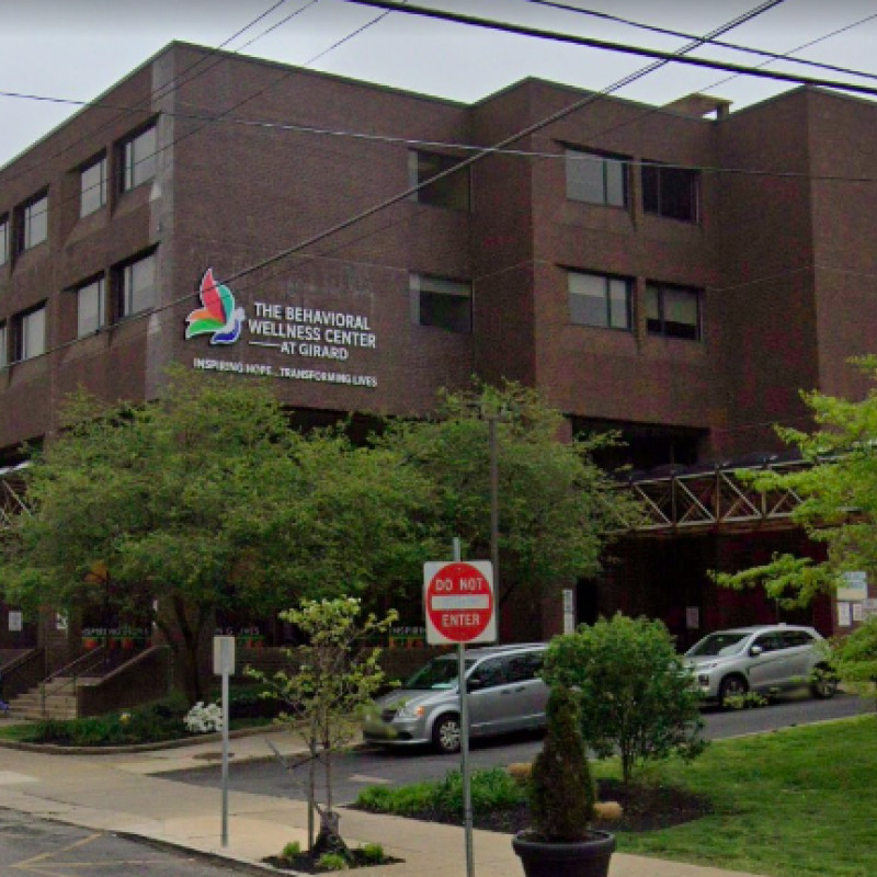 A brown brick building that says "The Behavioral Wellness Center at Girard" with green trees and some parked cars in the foreground