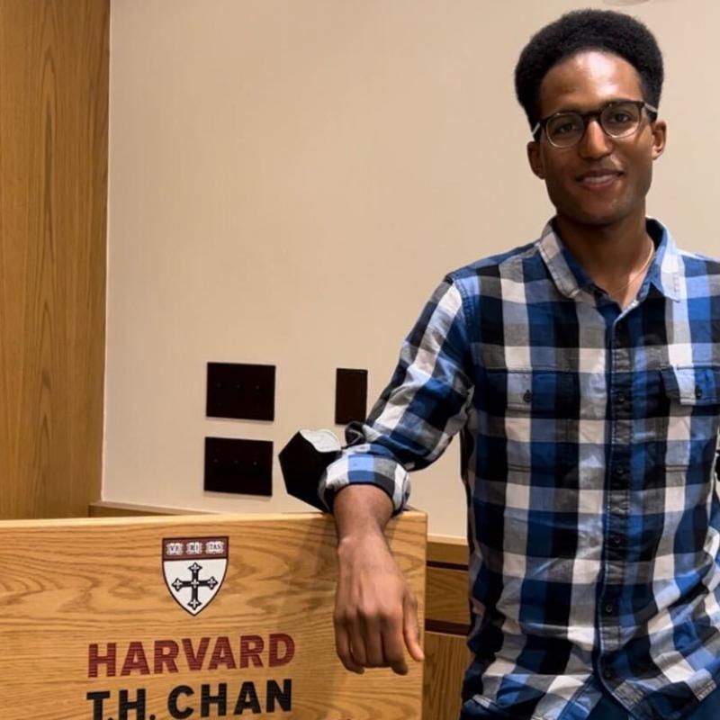 Spencer stands to the right of the frame, smiling at the camera. He is standing next to the podium showing the words "Harvard T.H. Chan School of Public Health"