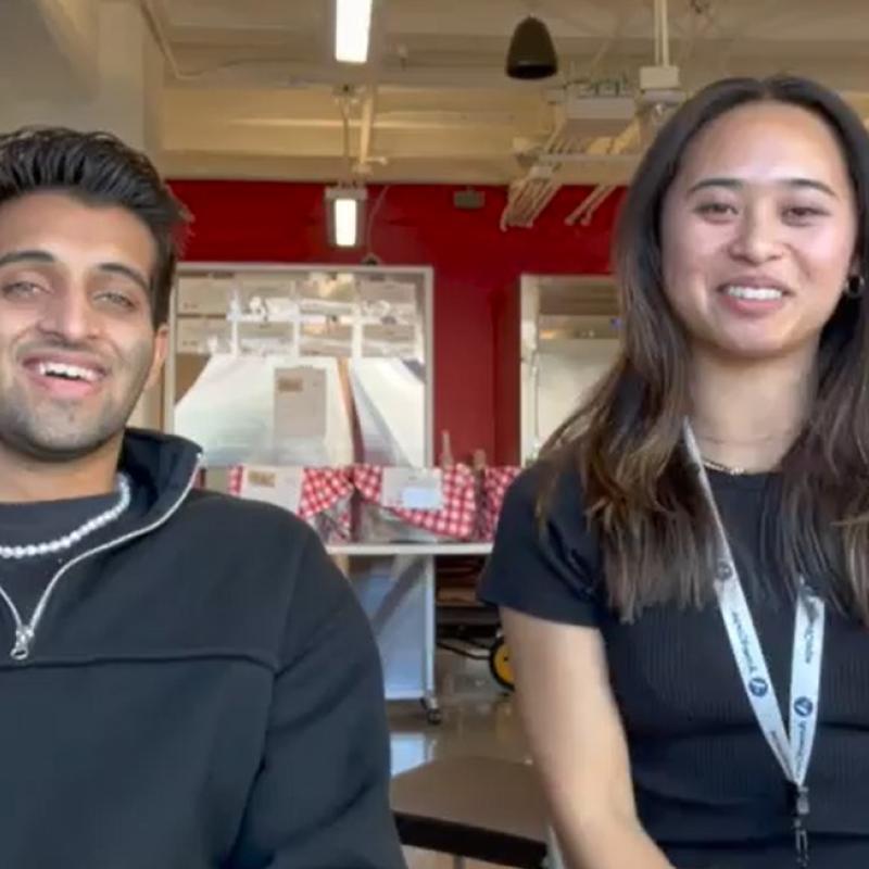 Abhi (left) and Laurel (right) sit in front of the camera, facing the camera and smiling. 