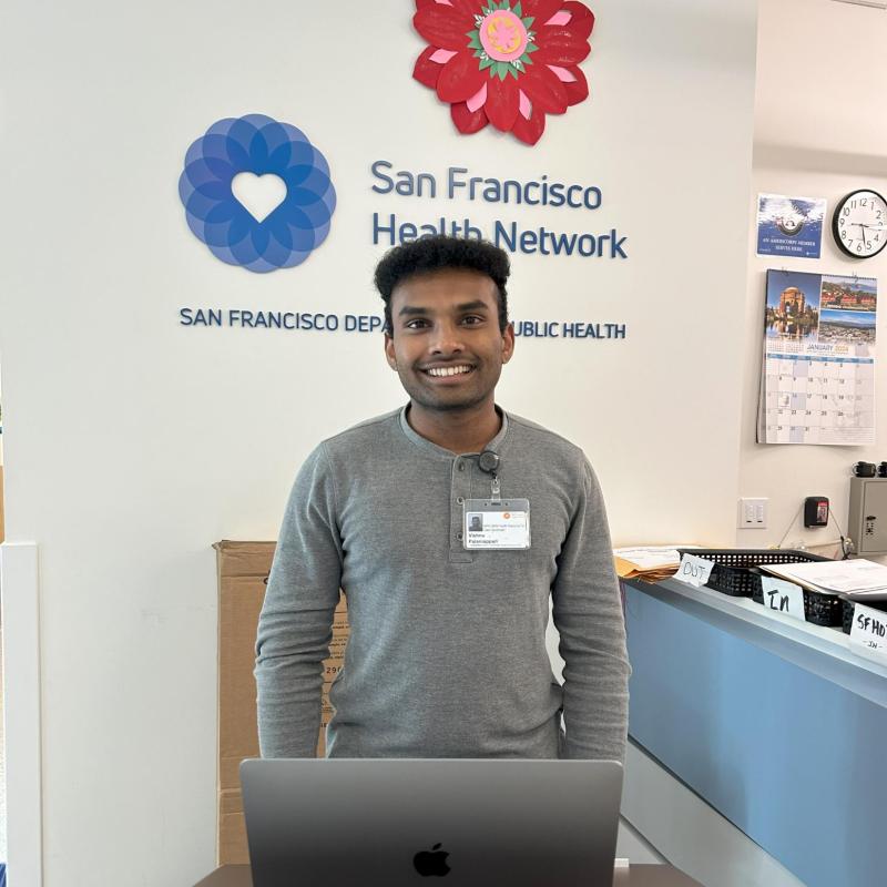 Vishnu is standing in the middle of the frame, facing the camera. He is standing behind a podium and has his laptop set on the podium in front of him. Behind him is the San Francisco Health Network logo. 
