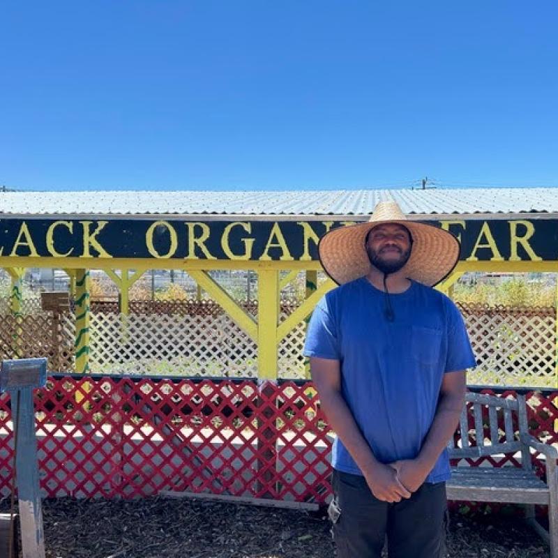 Faheem Carter stands in the center-right of the frame. Behind him is a farm shed with the sign "Black Organic Farm" painted onto the wooden overhang. 