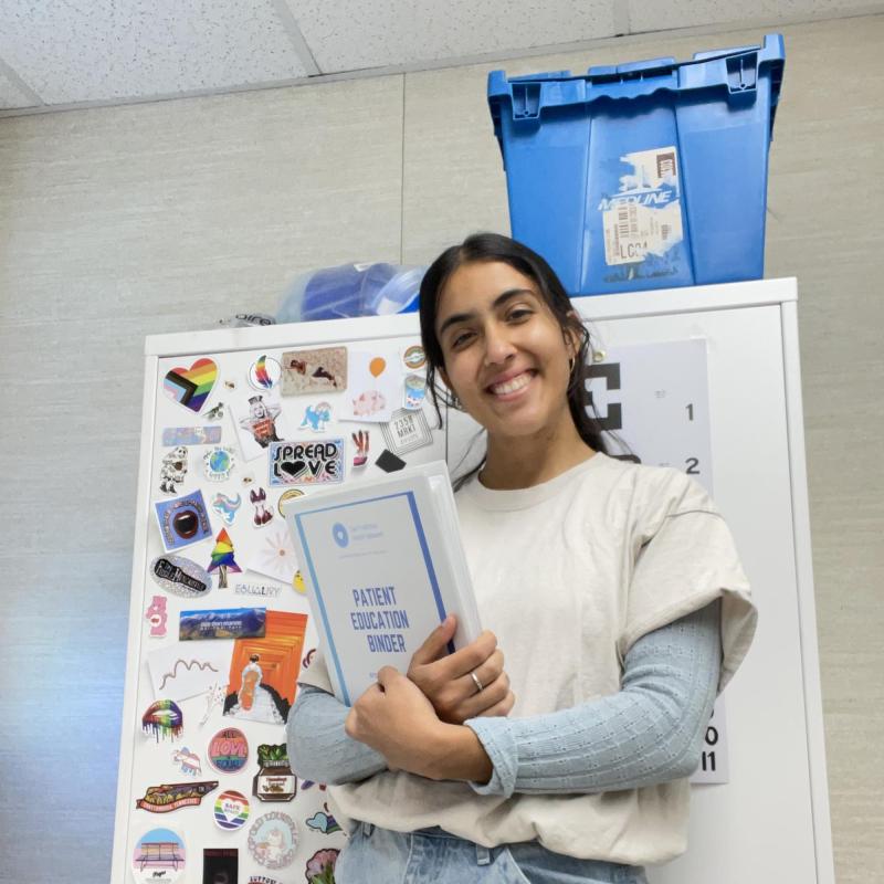 Anjalie is standing in the center of the frame, holding a binder. The cover of the binder reads "Patient Education Binder."