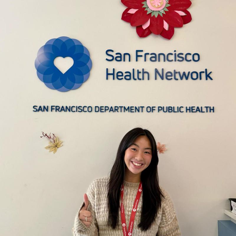Sam stands in the center of the frame, facing the camera and smiling. Behind her is the logo for the San Francisco Health Network.