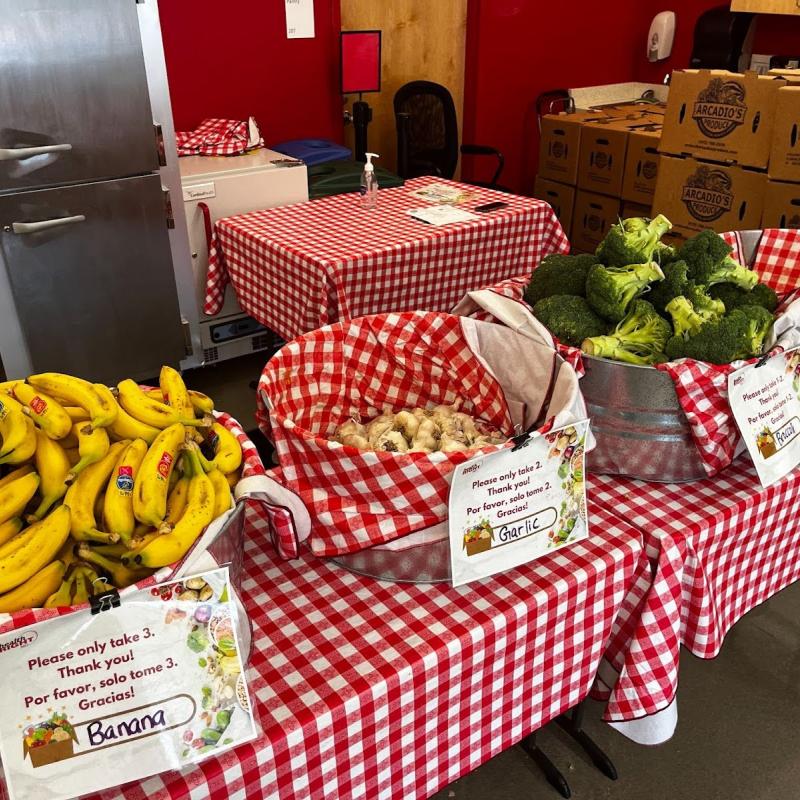 A table is covered with a checkered red and white tablecloth. On top of the table are baskets of fresh produce, including bananas, garlic, broccoli, and onions.